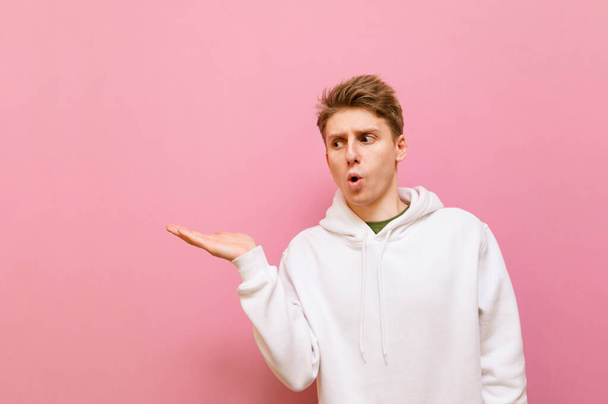 Funny guy stands on a pink background and looks to the side holding a copy space with a surprised face. Shocked young man holding an empty seat on his hand, Isolated. - Fotografie, Obrázek