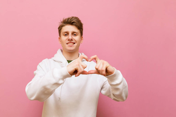 Ein hübscher junger Mann in weißen Kleidern zeigt eine Herzensgeste, blickt in die Kamera und lächelt auf rosa Hintergrund. Netter junger Mann zeigt Herz und posiert vor der Kamera. 14. Februar. Kopierraum - Foto, Bild