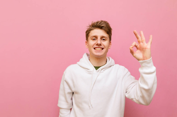 Positive guy in white hoodie stands on pink background, shows gesture Ok, looks into camera and smiles. Happy teenager in casual clothes showing thumbs up OK gesture isolated on pink. Copy space - Photo, Image