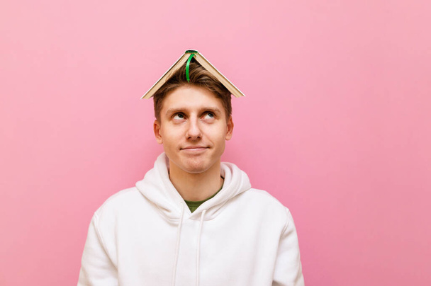 Funny guy student with a book on his head isolated on a pink background, looks away with a smile on his face and smiles. Cute young man holding a book on his head and looking away at copy space - Zdjęcie, obraz