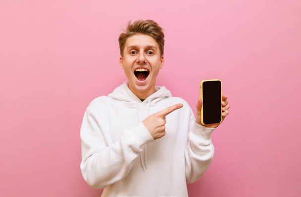 Щасливий молодий чоловік стоїть на рожевому фоні зі смартфоном в руці на рожевому тлі, дивлячись в камеру з здивованим обличчям і показуючи палець на чорному екрані. Копіювати простір
 - Фото, зображення