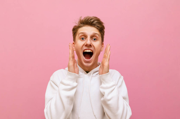 Closeup portrait of shocked guy in white hoodie on pink background looking into camera with mouth open and shouting with joy. Emotional young man yelling WOW, isolated. - Photo, Image