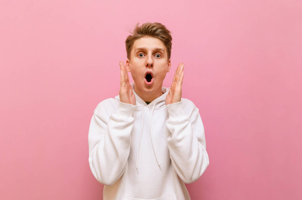 Shocked young man standing on a pink background, looking mad at the camera and putting his hands to his face. Portrait of surprised guy in white hoodie looking into camera with crazy face. Copy space - Photo, Image