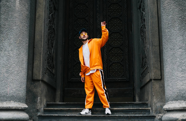 Street dancer in bright clothing dances modern hip hop on a background of dark architectureYoung man in an orange tracksuit shows hip hop performance on the street,standing on the steps old building - 写真・画像