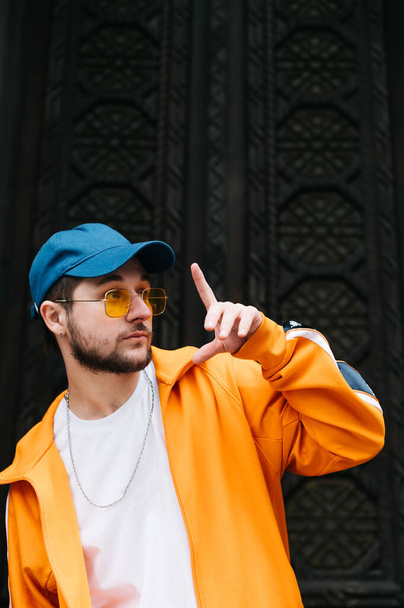 Portrait of a fashionable hip hop dancer on a dark background, wearing an orange tracksuit, looking away and posing at the camera. Stylish man in trendy street clothes on dark wall background, street - Foto, afbeelding