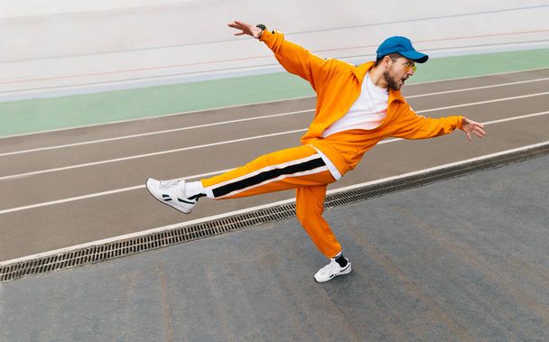 Stílusos hip-hop táncos bemutatja a teljesítményt a stadion pályán, visel egy narancssárga sportruházat öltöny és sapka. Táncosnőt képezni a játszótéren. Utcai tánc koncepció. Háttér. - Fotó, kép