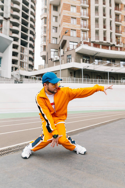明るい服のスタイリッシュな男は、スタジアムトラック上の近代的なダンスを踊り、オレンジのスポーツ衣装を身に着けています。屋外の遊び場でヒップホップダンサーのトレーニングの肖像画。ストリートダンス。垂直 - 写真・画像