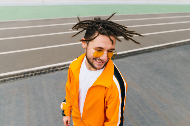 Uśmiechnięty facet w dredach i pomarańczowym dresie stoi na stadionie i uśmiecha się w okularach przeciwsłonecznych. Portret autentycznego młodego sportowca w okularach przeciwsłonecznych. - Zdjęcie, obraz