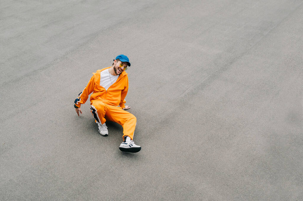 Photo of happy hip hop dancer in stylish bright streetwear sitting on asphalted playground. Cheerful young man dancing break dance, isolated on gray asphalt background. Background. Copy space - Fotó, kép