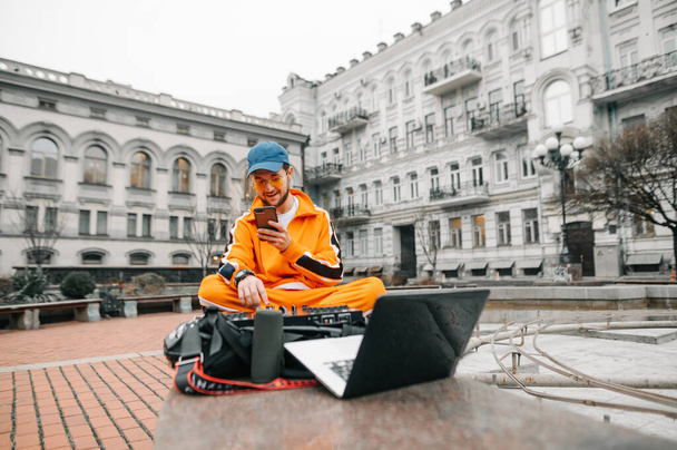Sokakta elinde bir kumanda, müzik sütunu ve dizüstü bilgisayarla oturan şık bir genç adam DJ seti çalıyor. Bir DJ 'in portresi kaldırımda oturmuş müzik karıştırıcısı ve dizüstü bilgisayarla müziği karıştırıyor.. - Fotoğraf, Görsel