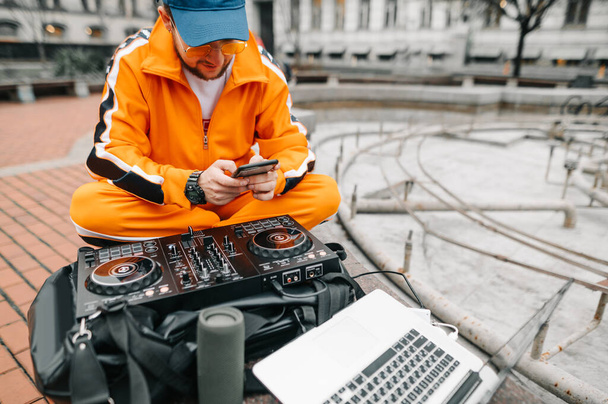Nahaufnahme eines stilvollen DJs, der mit Controller, Laptop, Musiklautsprecher draußen sitzt, Musik mixt und ein Smartphone benutzt. Street-DJ spielt Musik im Freien. Kopierraum - Foto, Bild
