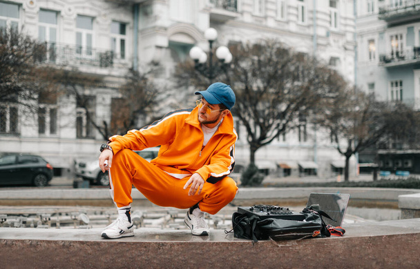 Modieuze jongeman in casual kleding zit met laptop, muziekmixer en luidspreker op straat en speelt muziek. DJ speelt muziek op een controller op straat en kijkt weg met een serieus gezicht - Foto, afbeelding