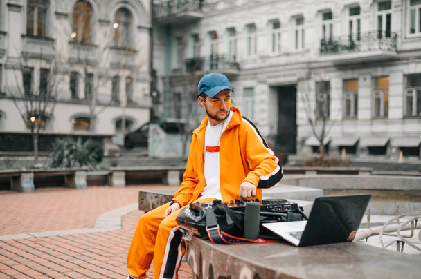 Stylowy młody człowiek siedzący na ulicy z kontrolerem, kolumną muzyczną i laptopem grającym zestaw DJ-ów. Portret DJ-a siedzącego przy krawężniku z mikserem muzycznym i laptopem, mieszającego muzykę. - Zdjęcie, obraz