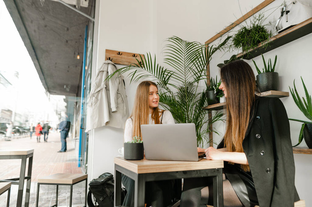 Twee jonge meisjes in formele kleding zitten in een gezellig café met een laptop en bespreken zaken met een glimlach op hun gezicht. Jonge zakenvrouwen kletsen in een gezellig café met een kop koffie. - Foto, afbeelding