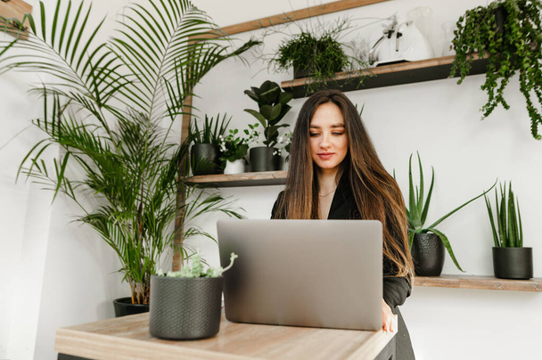 Привлекательная деловая женщина с темными волосами, носит черную куртку, работает на ноутбуке в уютном кафе, смотрит на экран с улыбкой на лице.
 - Фото, изображение
