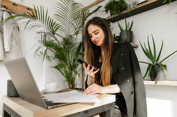 Portret van een positief kantoormeisje in een jasje zittend aan een koffiepauze en werkend op een laptop, kijkend naar smartphone met een glimlach op haar gezicht.Zakenvrouw gebruikt smartphone en laptop in café - Foto, afbeelding