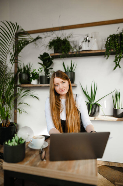 Schattig freelancer meisje met laptop werkend in een gezellig café, kijkend in de camera en lachend. Aantrekkelijk studentmeisje gebruikt het internet op een laptop in een stijlvolle coffeeshop. Verticaal - Foto, afbeelding