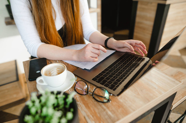 Een close-up foto. Studenten meisje zit met laptop en papieren in cafe met kopje koffie en studeren. Lady freelancer werkt op de laptop en drinkt koffie in een koffiewinkel.Werken in een café. Achtergrond - Foto, afbeelding