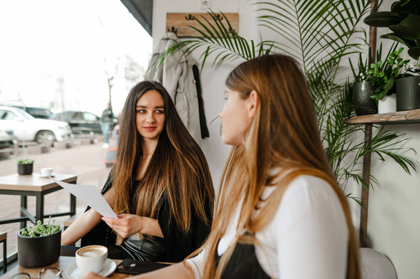 Chica atractiva en ropa formal en una reunión de negocios en un café hablando y discutiendo el proyecto. Las compañeras están sentadas en una cafetería tomando una taza de café y hablando con sonrisas
. - Foto, imagen