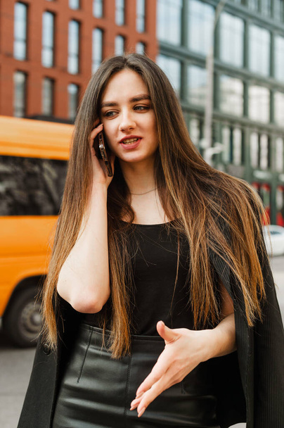 Portrait of a businesswoman in dark formal wear talking on the street and actively gesturing when explaining. Beautiful young girl office worker in dark roe clothes clothes calling on smartphone. - Photo, image