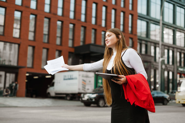 Zaneprázdněná studentka stojí s laptopem a papíry v rukou a chytí gesto ruky taxikáře s úsměvem na tváři. Pozitivní dívka ve formálním oblečení zastaví taxi na ulici metropole. - Fotografie, Obrázek