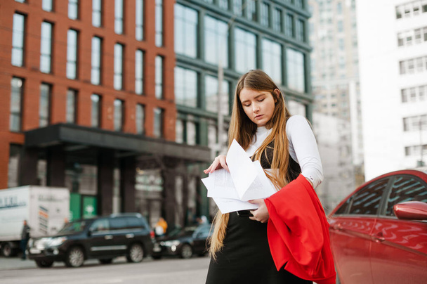 Portré elfoglalt diáklány áll a szabadban városkép háttér laptop és jegyzetek a kezében, zavaros keresi a megfelelő papírt. A kocka lány az egyetemre tanul.. - Fotó, kép