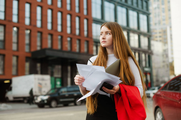 Porträt eines netten Studentenmädchens, das mit Laptop, Smartphone, Notizen in der Hand auf der Straße einer Großstadt steht und mit ernstem Gesicht wegschaut. - Foto, Bild