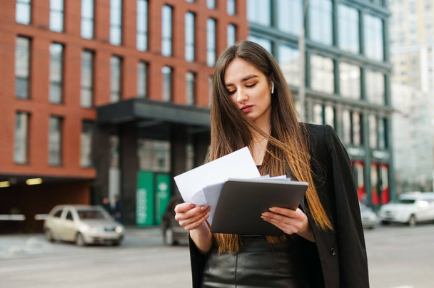 Business girl in wireless headphones stands with laptop and business papers against a backdrop of a street landscape and reads information on documents with a serious face. Business people concept. - Valokuva, kuva