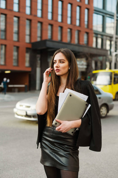 Vertikales Porträt einer eleganten Geschäftsfrau in formeller Kleidung, die mit Laptop und Dokumenten in der Hand am Straßenrand steht, wegschaut und auf ein Taxi wartet, kommuniziert über drahtlose Kopfhörer - Foto, Bild