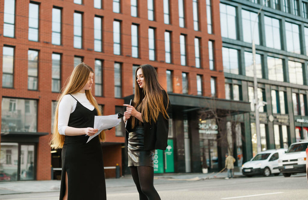 Dwie biznesmenki stoją na ulicy z laptopem i dokumentami biznesowymi w ręku i rozmawiają.Pracownice biurowe dziewczyn dyskutują o pracy na tle miejskiego krajobrazu. - Zdjęcie, obraz