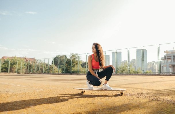 Attraktives hispanisches Mädchen in trendiger Streetwear sitzt auf einem Longboard auf dem Basketballfeld und blickt mit ernstem Gesicht weg. Street Fashion Mädchen Skater posiert auf Longboard. - Foto, Bild