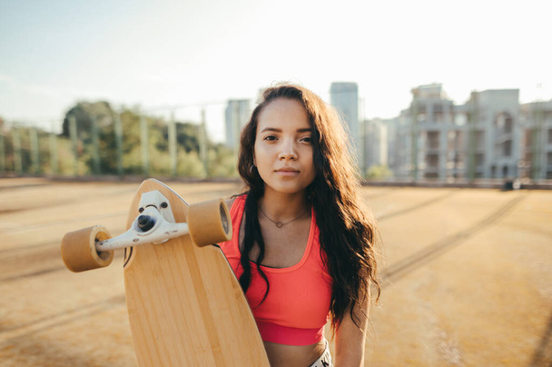 Closeup portrait of attractive hispanic girl in casual clothes standing on the street with longboard in her hands, looking into the camera with a smile on her face. Street style. Skateboard concept. - Photo, Image