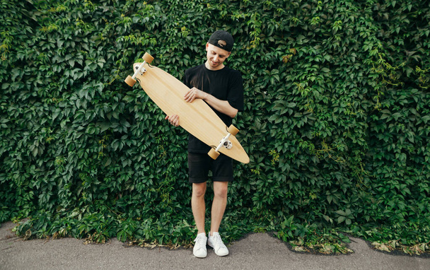 Young skateboarder in dark clothing stands against the background of a wall with a vine, holds a longboard in hands and happy looks down. Portrait of teenager with longboard on green wall background - Photo, Image