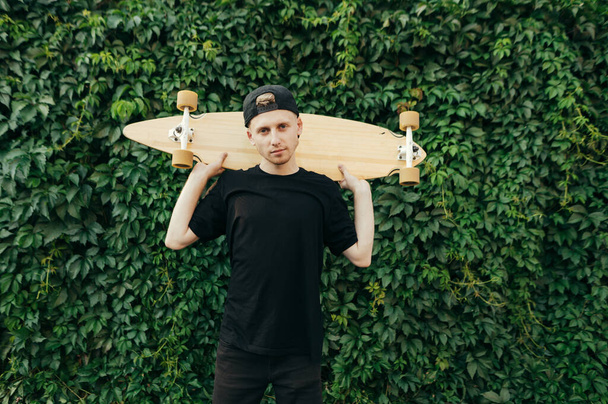Portrait of a handsome young man in casual clothes and cap posing with a longboard in his hands on a green background with a vine, looking into the camera with a serious face. Copy space - 写真・画像