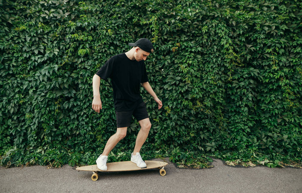 Young man rides logboard on wall background with ivy plant, side photo. Teenager walking on a skateboard on a street town on a background of a wall with a vine. Copy space - Zdjęcie, obraz