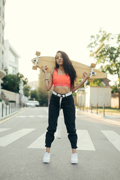 Portrait of attractive Hispanic woman in trendy streetwear posing at camera with skateboard in hands. Beautiful street girl with a longboard behind her shoulders stands on the road - Photo, Image