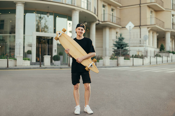 Full length portrait of teenager boy standing with longboard in hands outdoors, looking at camera and smiling. Young man in dark clothes posing at camera with skate in hands. Copy space - Photo, Image