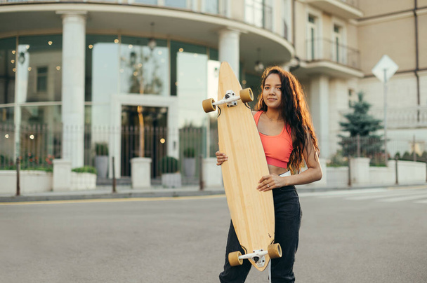A portrait of a smiling girl in stylish street clothes standing on the street at sunset with a longboard in her hands, looking into the camera with a happy face. Hispanic girl skater posing. - Photo, Image