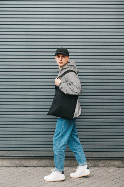 Portrét módního mladého muže v pouličních šatech stojí na pozadí šedé stěny s černou eko taškou v ruce a dívá se do kamery s vážnou tváří. Svisle - Fotografie, Obrázek