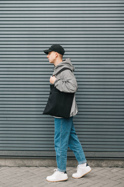 Modaya uygun bir sokak adamı omuzlarında eko çanta, uzun bir portre ile gri bir duvarın arka planında yürüyor. Dikey. Çevre dostu - Fotoğraf, Görsel