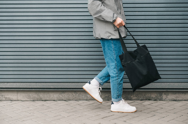 Καλλιεργημένη φωτογραφία του πάτου ενός νεαρού άνδρα με κομψά ρούχα που περπατάει στο φόντο ενός γκρίζου τοίχου με μια μαύρη οικολογική τσάντα στο χέρι του. Οικολογικό. Επαναχρησιμοποιήσιμη τσάντα στο χέρι του νεαρού. Αντιγραφή χώρου - Φωτογραφία, εικόνα