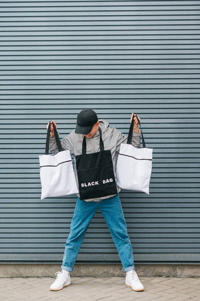 Smiling eco friendly guy stands against a gray wall with 3 reusable shopping bags looking down. Fashionable photo of a young man in streetwear with eco bags in his hands. Vertical - Foto, afbeelding