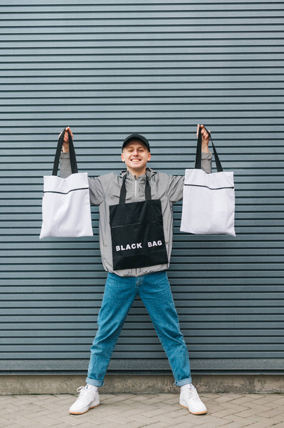 Happy young man in stylish casual clothes stands with 3 eco bags in his hands and poses at the camera with a smile on his face. Eco friendly guy in trendy streetwear shows reusable fabric bags. - Φωτογραφία, εικόνα