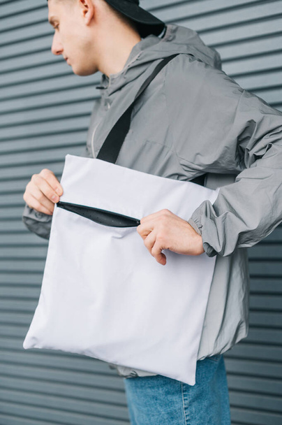 Closeup photo, stylish young man opening white eco bag with lightning. Vertical photo. Background. Advertising stylish eco bags. - Фото, зображення