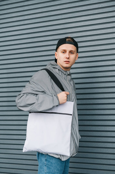 Portrait of attractive young man in stylish streetwear with white eco bag on shoulder posing on gray background with serious face. Guy with stylish reusable eco bag. Vertical. - Photo, Image
