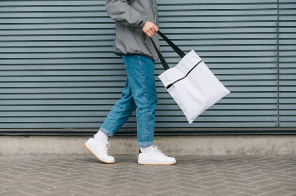 灰色の背景、足やエコバッグを閉じる上で再利用可能なショッピングバッグと歩いて通りの服でトリミングされた写真、スタイリッシュな若い男。コピースペース - 写真・画像