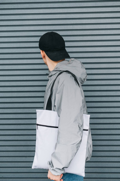 Stylish guy in a reflective jacket and cap stands against a gray wall with a trendy white eco bag turned away from the wall. Vertical photo. Background. - Fotografie, Obrázek