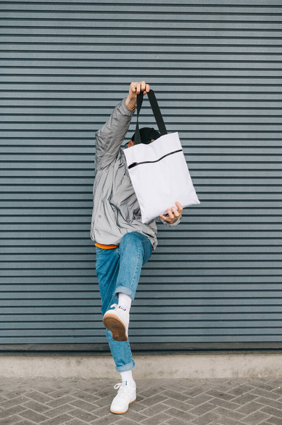 Funny young man in trendy streetwear having fun with eco bag in his hands on gray wall background, vertical photo. An expressive guy holds a reusable shopping bag in his hand and lifts his leg. - Photo, image
