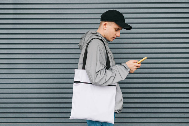 Młody człowiek w stylowych, ulicznych ciuchach i z białą eko torbą na ramieniu stoi na szarym tle i używa smartfona.Facet trzymający smartfona i niosący torbę na zakupy wielokrotnego użytku. Przestrzeń kopiowania - Zdjęcie, obraz