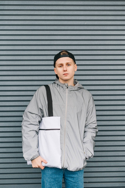 Portrait of a fashionable guy in street clothes and a cap stands against a background of a gray wall with a reusable eco bag on his shoulders, looking into the camera with a serious face. Vertical - Foto, afbeelding